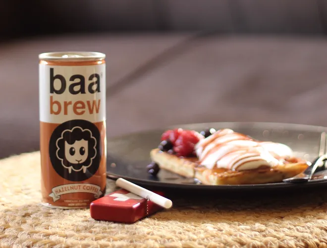 A Baa Brew hazelnut coffee can stands beside a red lighter with a cigarette and a plate of waffles topped with berries and whipped cream, all on a woven mat with a blurred sofa behind.
