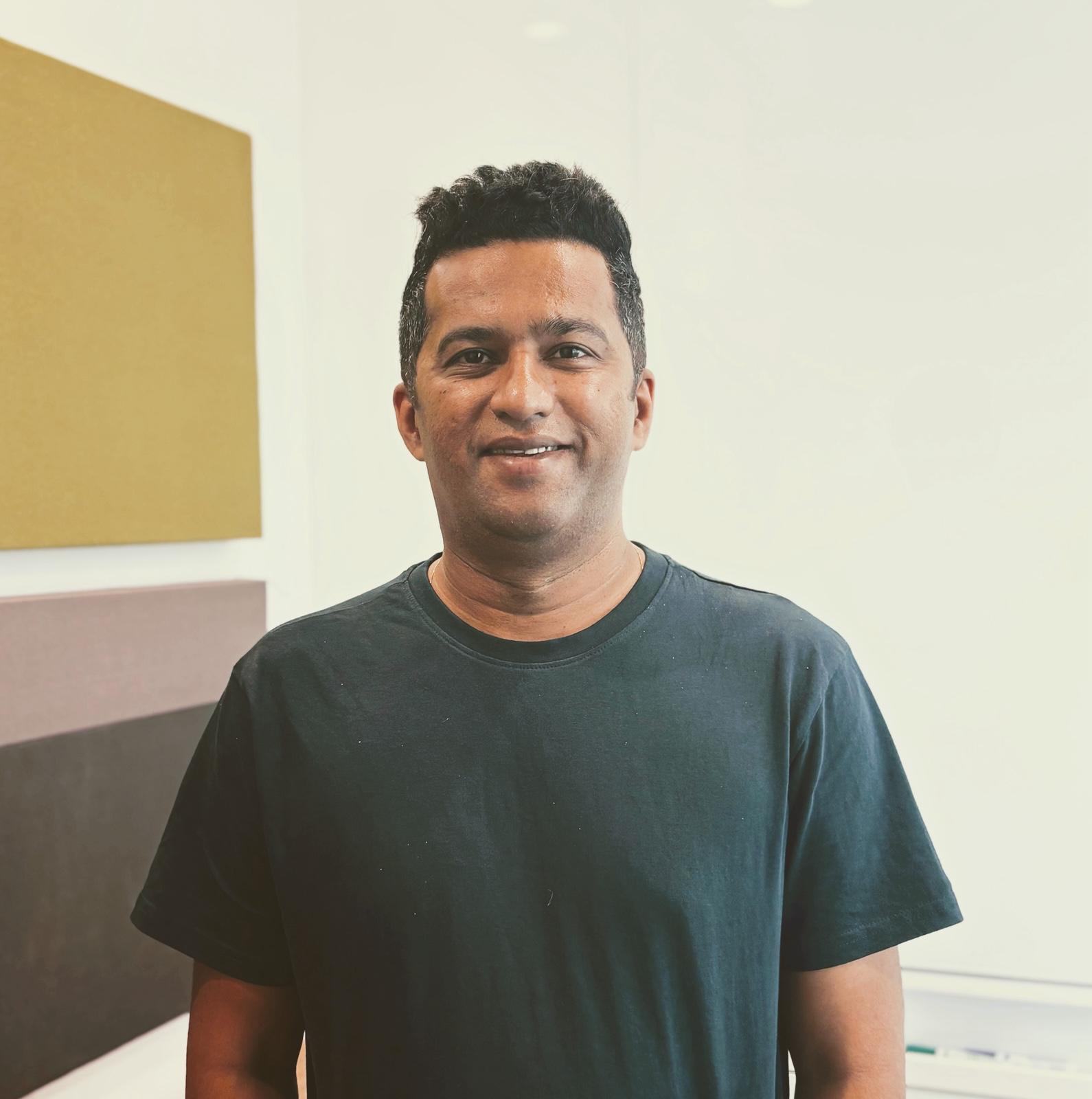 Smiling man in a dark t-shirt against a light wall with colored panels.