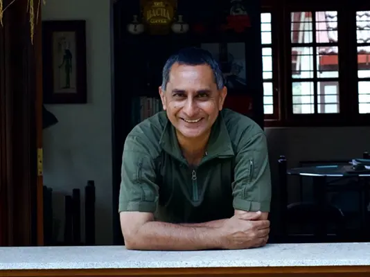 A smiling man in a dark green shirt leans on a counter indoors.