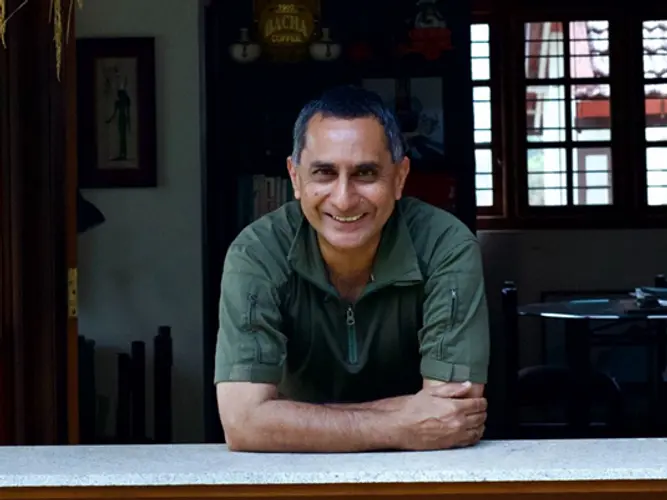 A smiling man in a dark green shirt leans on a counter indoors.