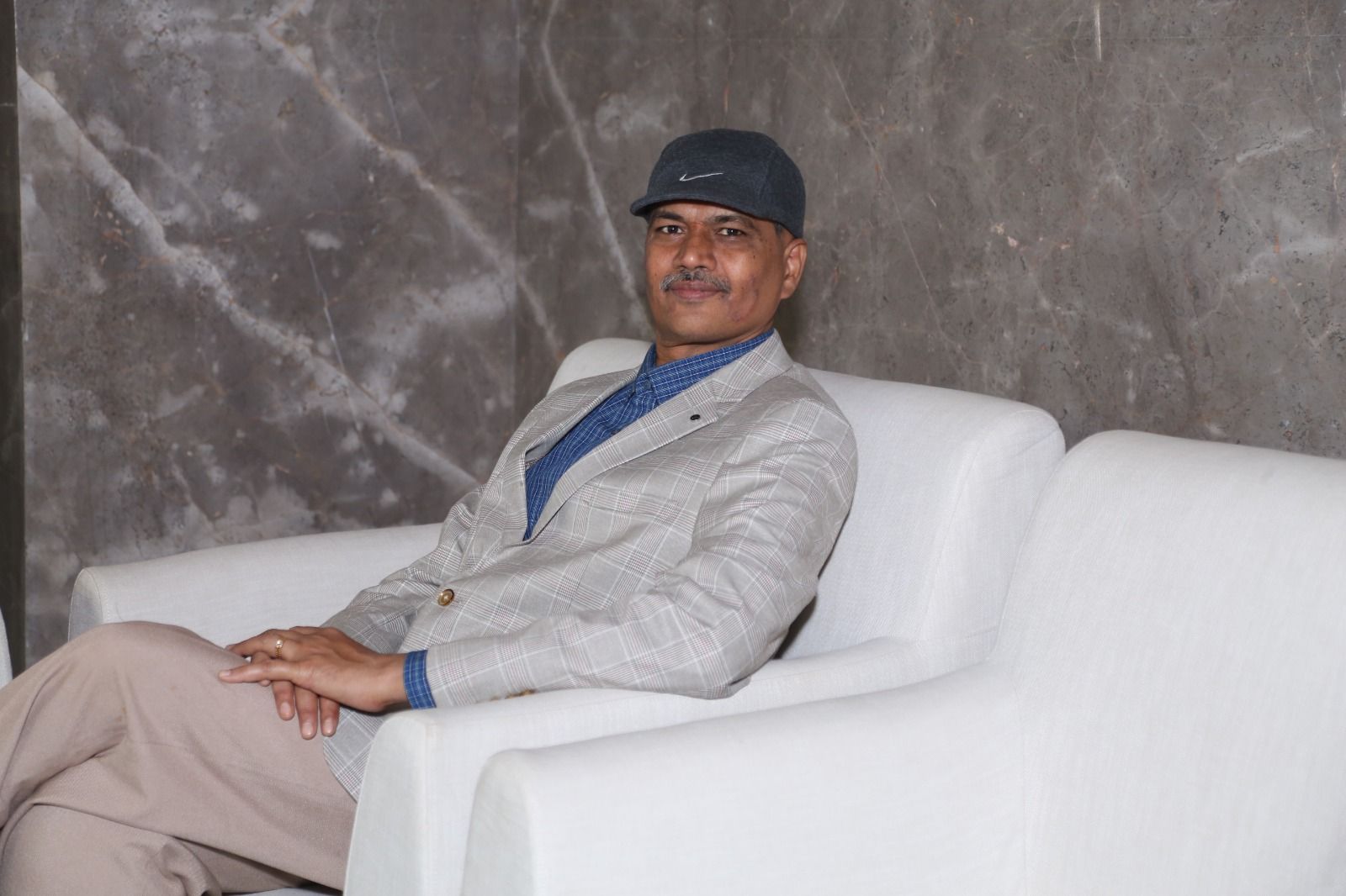 A man in a cap and blazer sits on a white sofa.