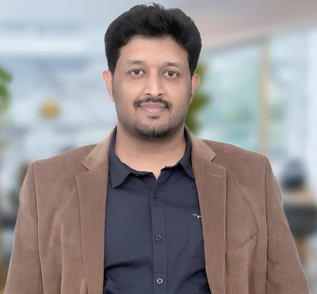 A man with dark hair and a beard, wearing a brown corduroy jacket over a dark shirt, looks at the viewer.