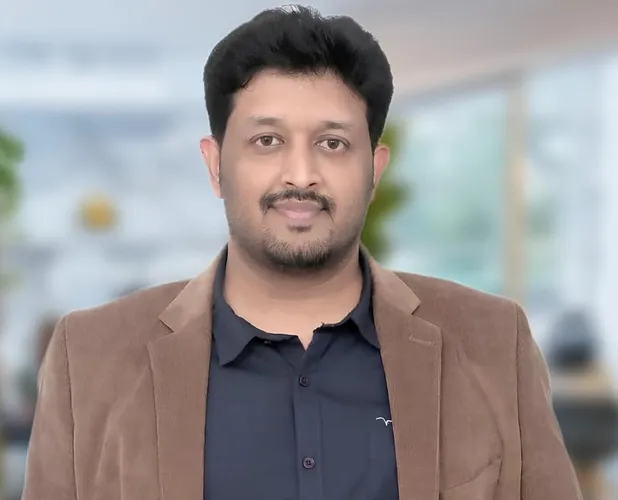 A man with dark hair and a beard, wearing a brown corduroy jacket over a dark shirt, looks at the viewer.