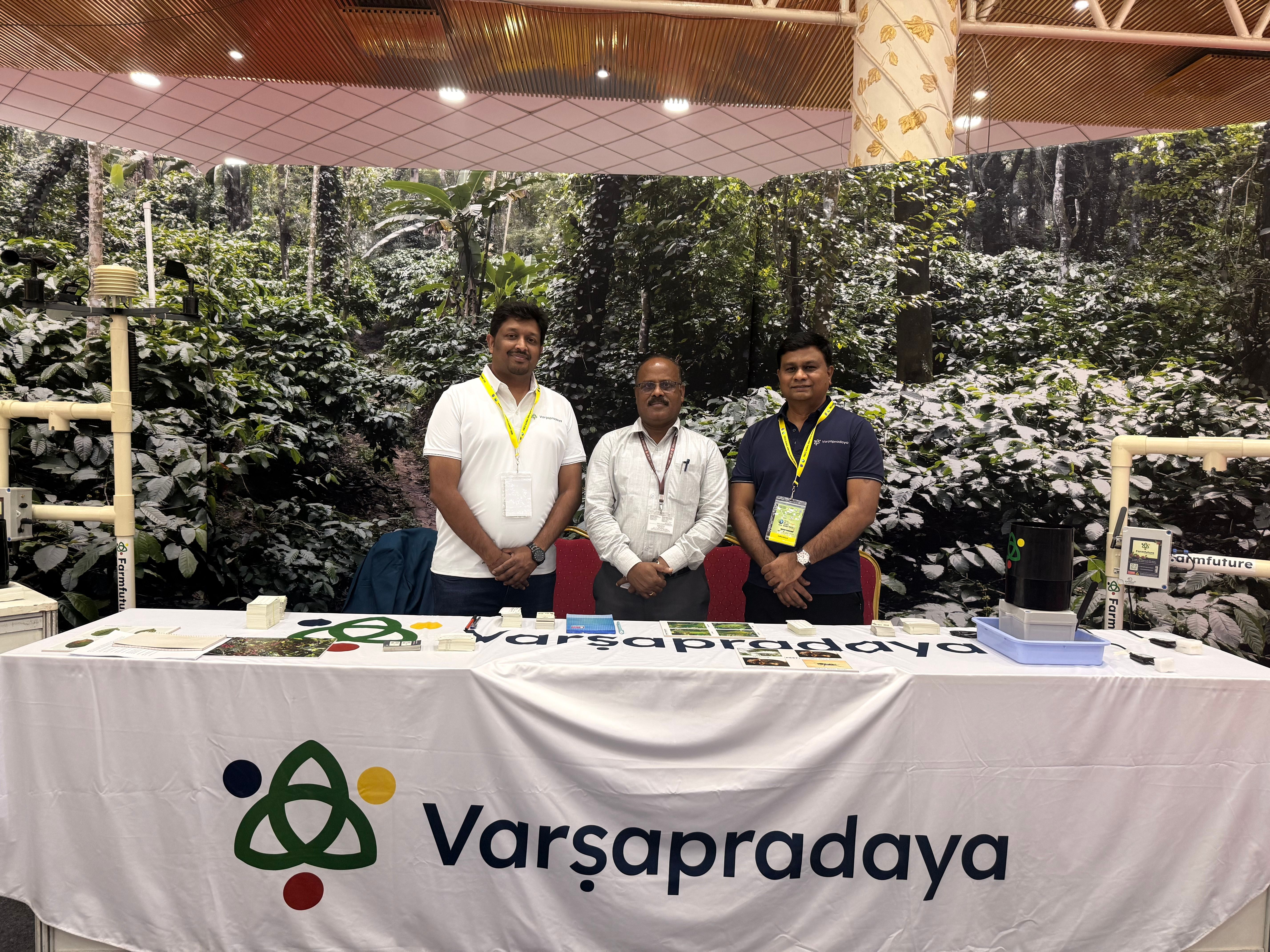 Three men standing behind the "Varsapradaya" booth with agricultural sensing equipment and a green plant backdrop.