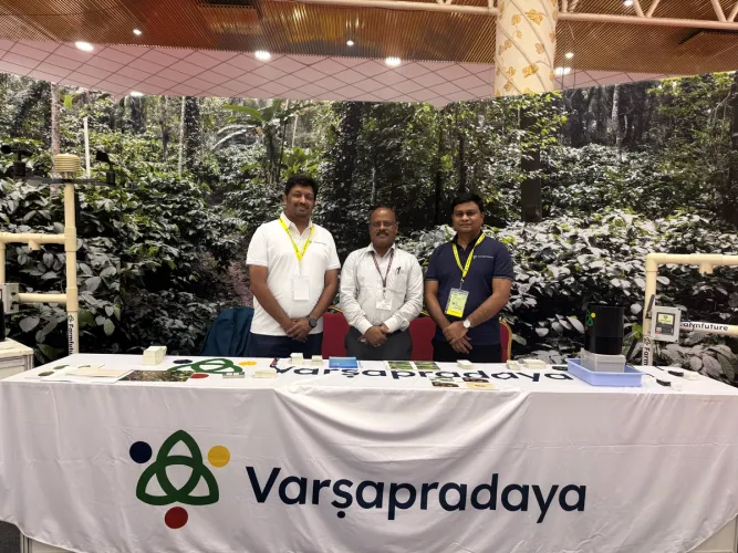 Three men standing behind the "Varsapradaya" booth with agricultural sensing equipment and a green plant backdrop.
