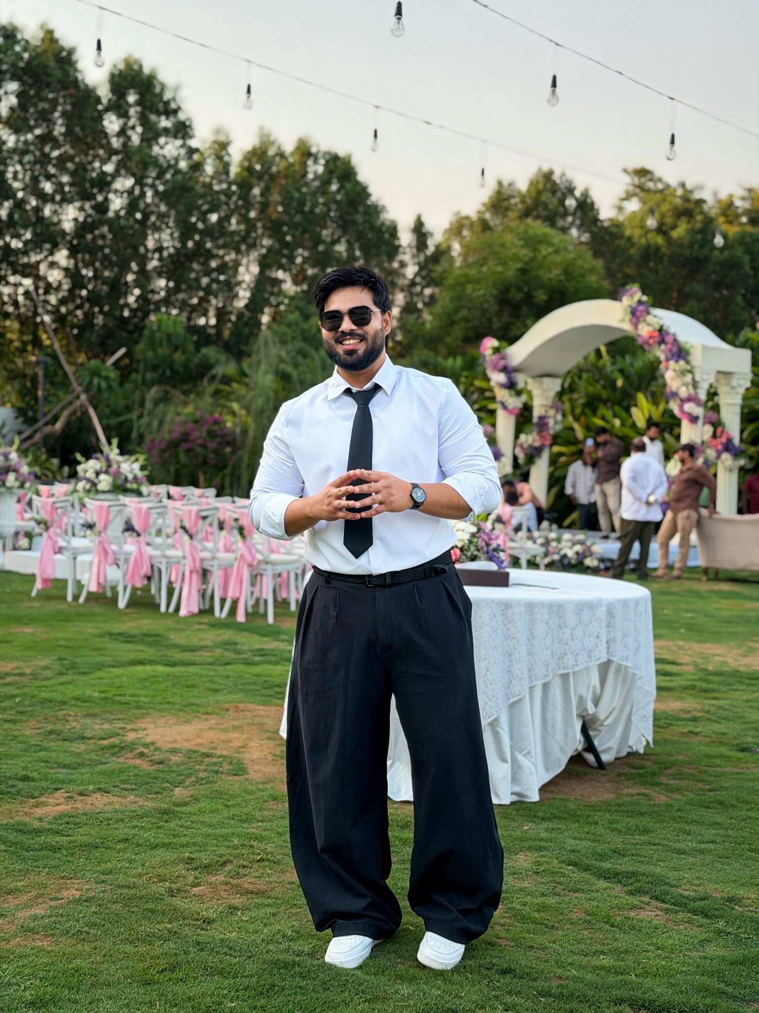 A smiling man in sunglasses, a white shirt, black tie, and wide-leg black pants stands on a lawn at an outdoor event.