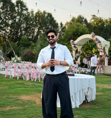 A smiling man in sunglasses, a white shirt, black tie, and wide-leg black pants stands on a lawn at an outdoor event.