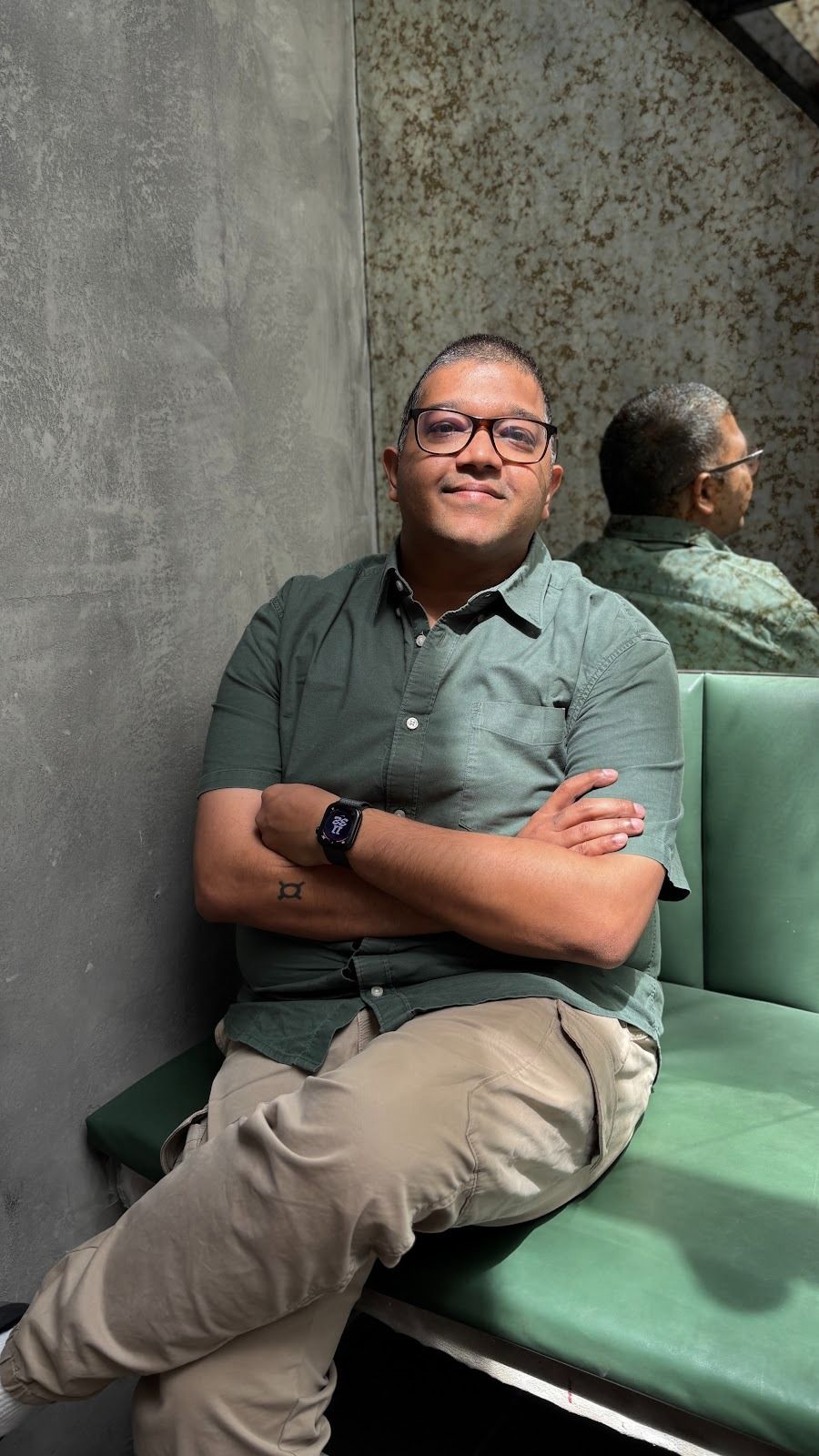 A man in glasses and a green shirt sits smiling with crossed arms on a green bench, his reflection visible in a mirror behind him.
