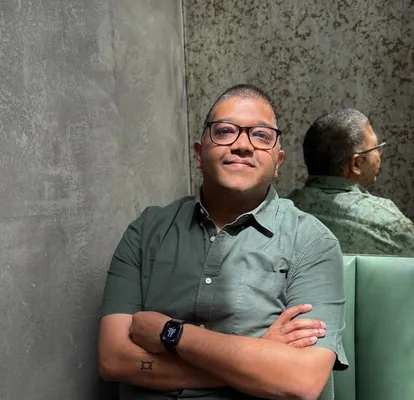 A man in glasses and a green shirt sits smiling with crossed arms on a green bench, his reflection visible in a mirror behind him.