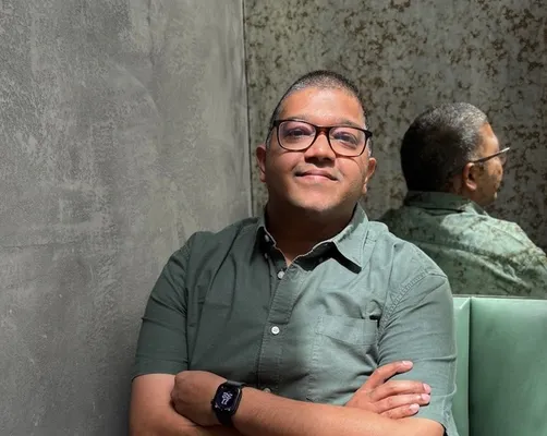 A man in glasses and a green shirt sits smiling with crossed arms on a green bench, his reflection visible in a mirror behind him.
