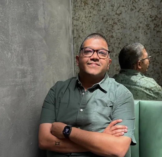 A man in glasses and a green shirt sits smiling with crossed arms on a green bench, his reflection visible in a mirror behind him.