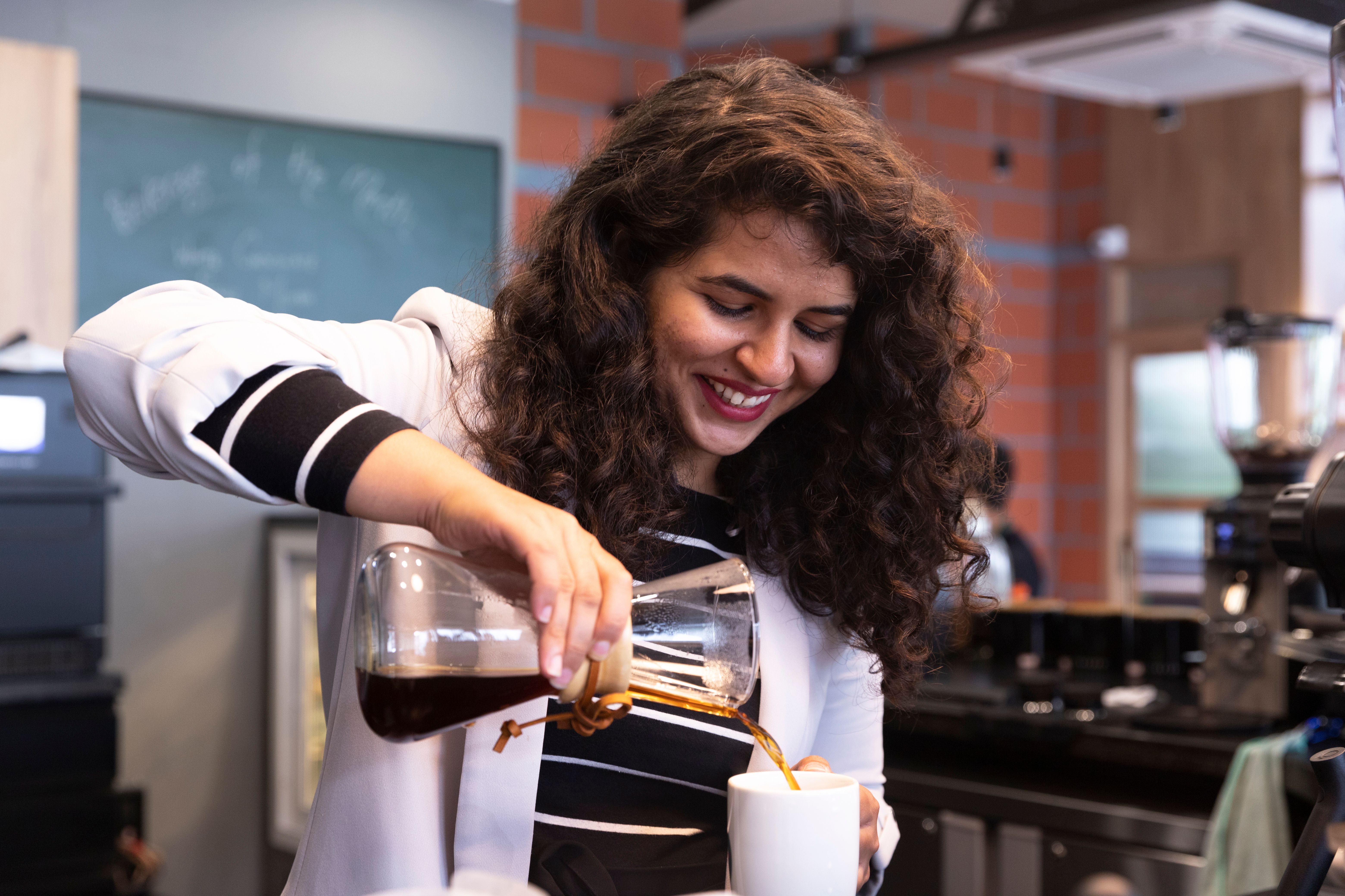 A smiling woman with curly hair pours coffee from a glass carafe into a white mug.
