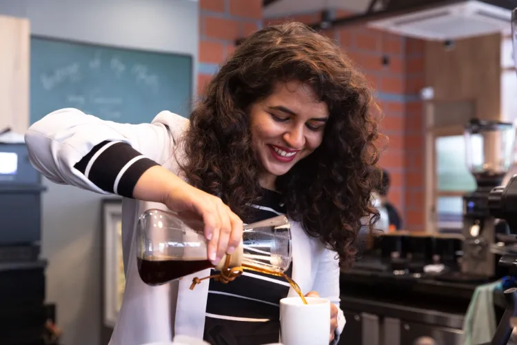 A smiling woman with curly hair pours coffee from a glass carafe into a white mug.