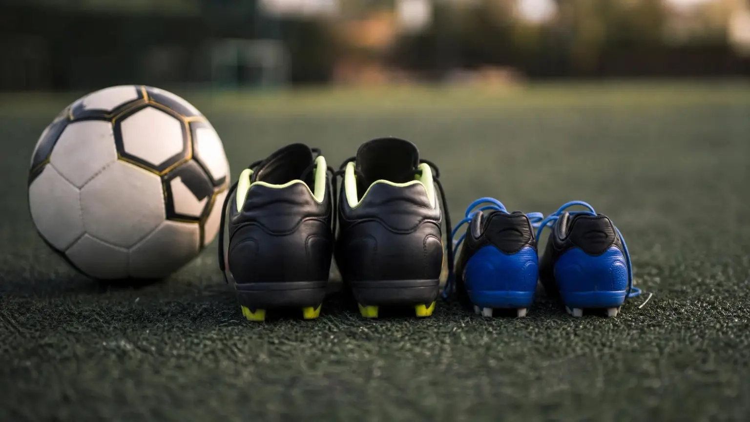 Two pairs of soccer cleats side by side on turf, one pair noticeably larger than the other, illustrating the size gap involved in playing up an age group in youth soccer