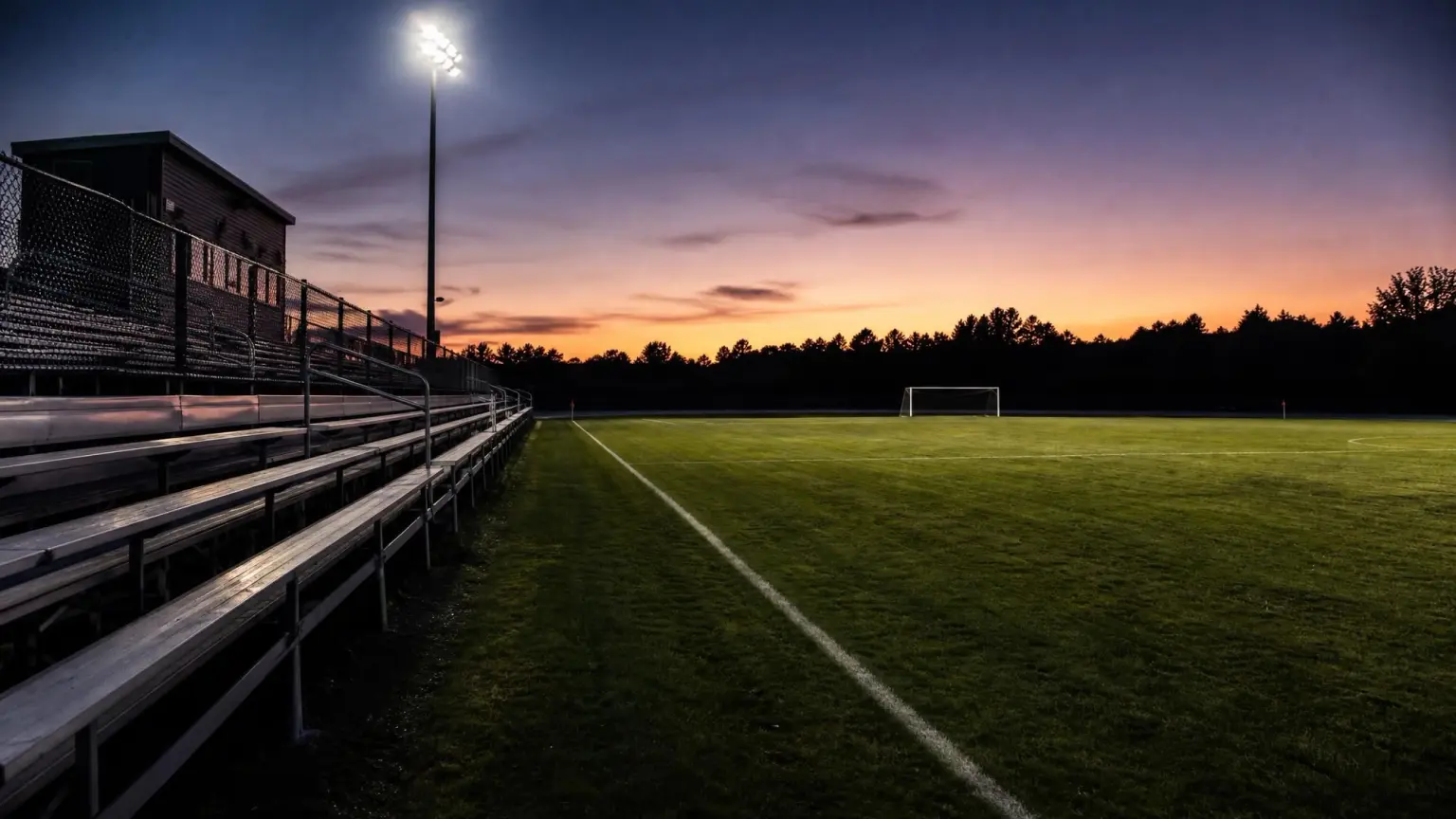 An empty high school soccer field at dusk with metal bleachers along the touchline and floodlights switched on, illustrating the club soccer vs high school soccer decision