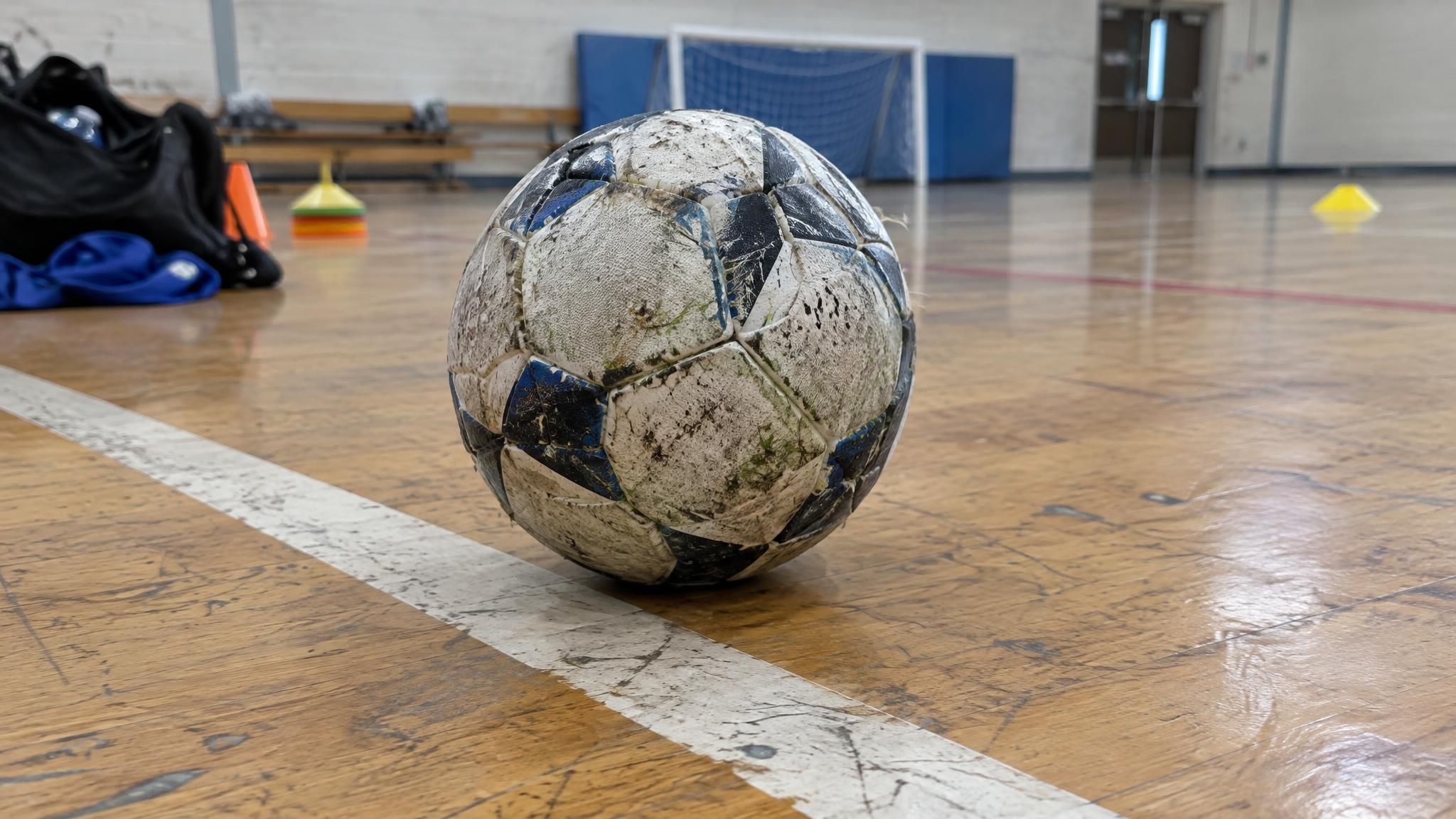 A low-bounce futsal ball resting on the wooden floor of a school gym beside a painted court line, ready for indoor soccer drills