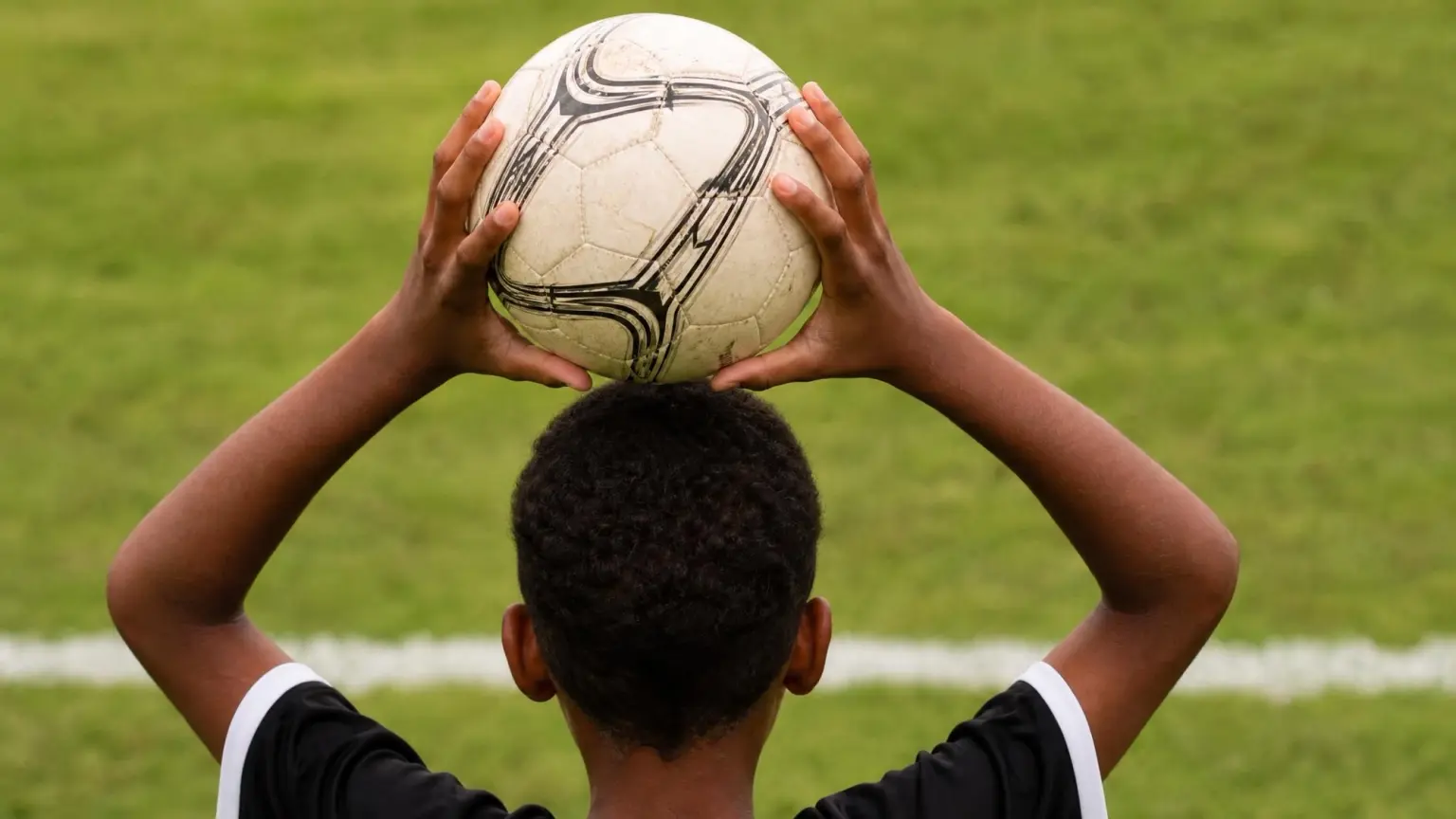 A player grips a soccer ball with both hands behind and above their head at the touchline, the position required by the soccer throw-in rules.