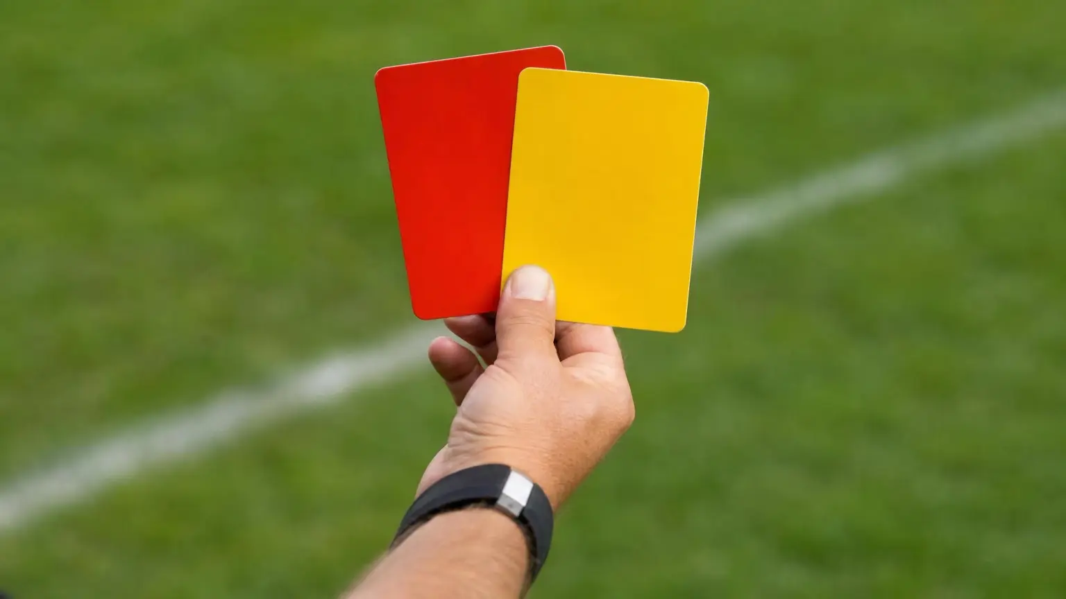 A referee's yellow and red cards held up against a blurred green pitch, illustrating yellow and red cards in soccer