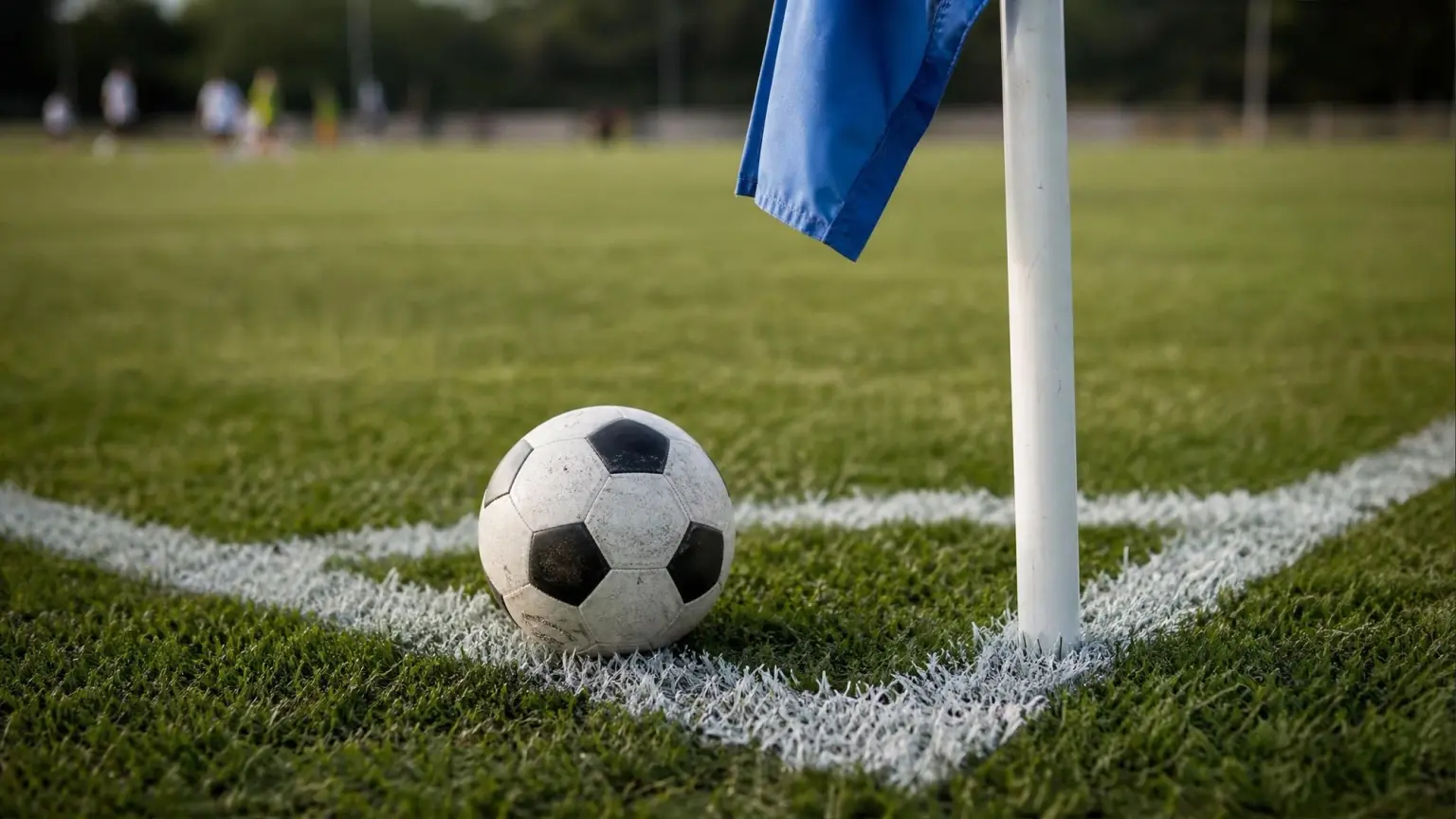 A soccer ball resting inside the painted corner arc of a grass pitch next to the corner flag, ready for a corner kick