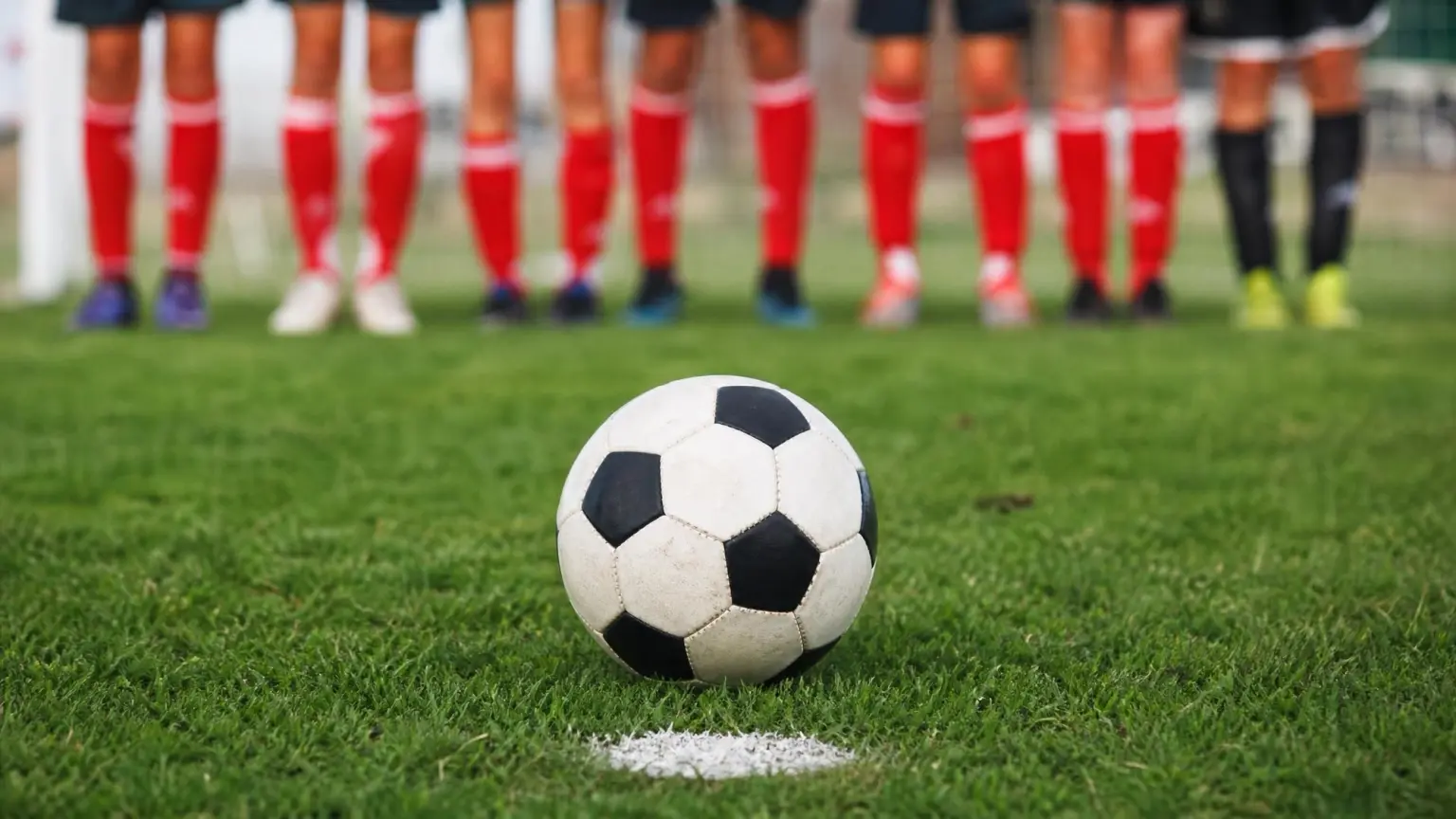 A soccer ball resting on the grass ready to be struck, with a blurred defensive wall behind it, illustrating a direct vs indirect free kick in soccer
