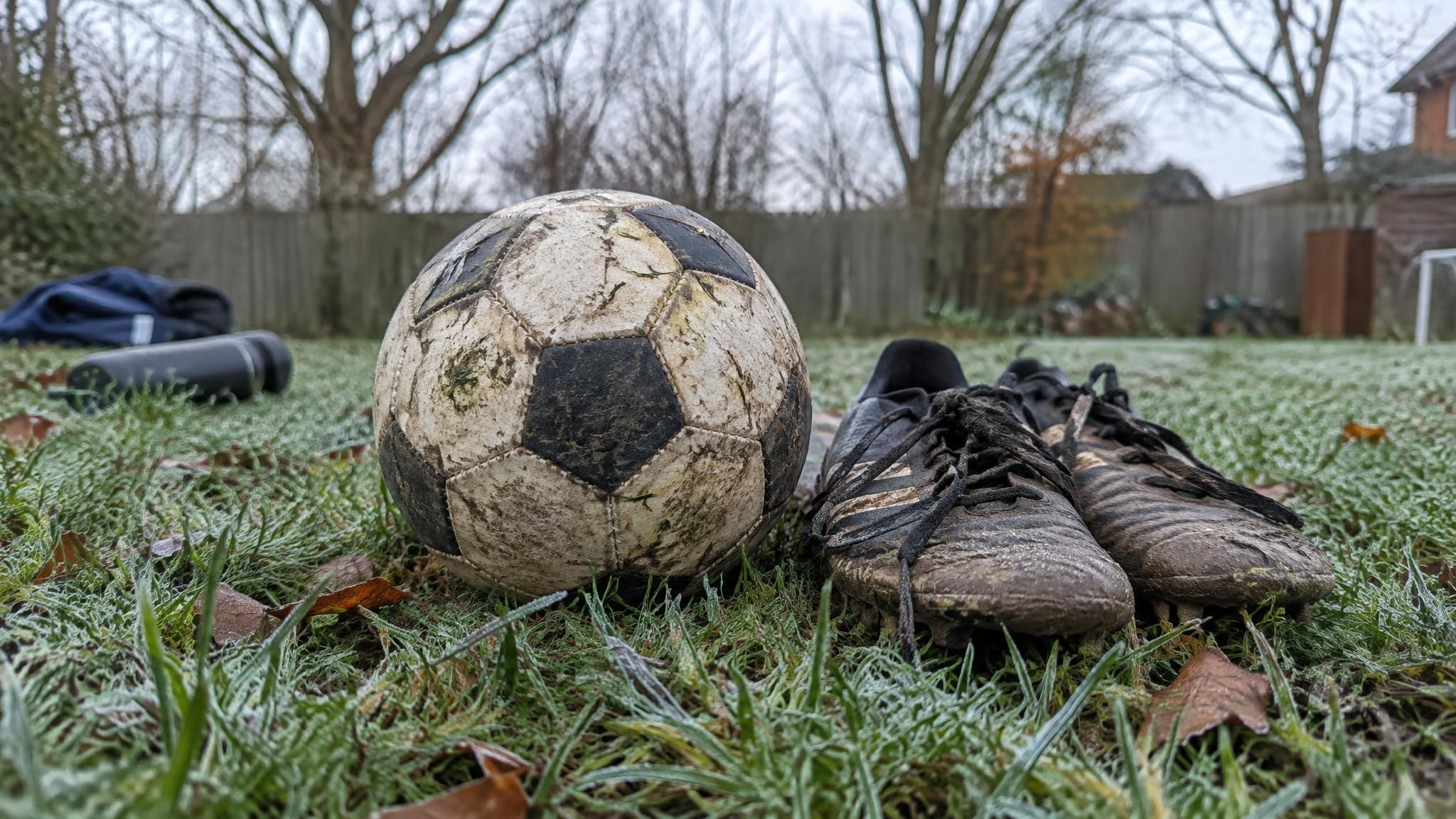 A worn soccer ball and muddy youth soccer cleats resting on frosty grass during the off-season