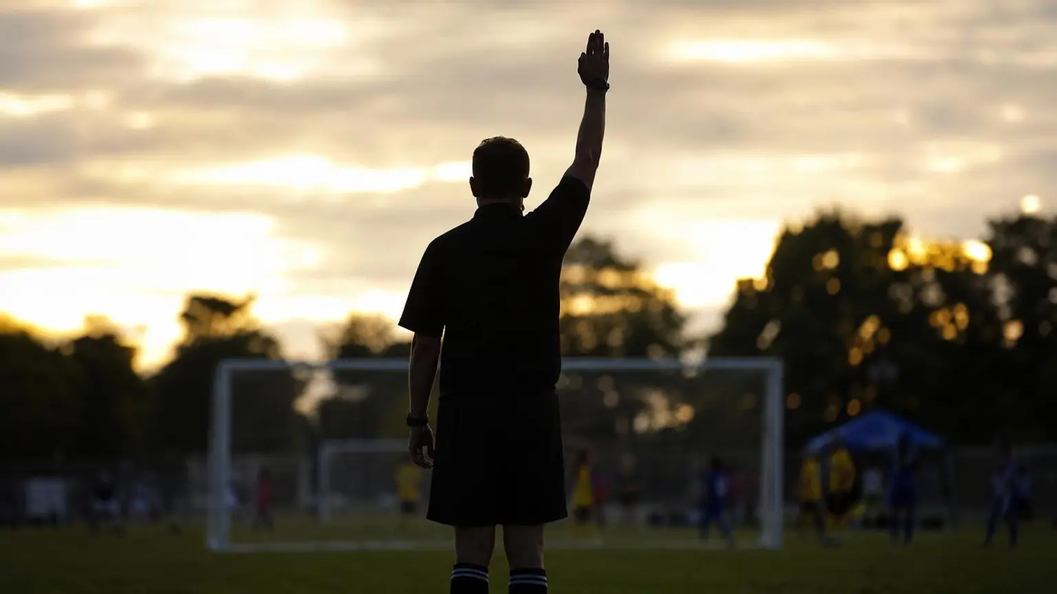Silhouette of a soccer referee on a youth pitch with one arm raised straight overhead, the standard soccer referee signal for an indirect free kick
