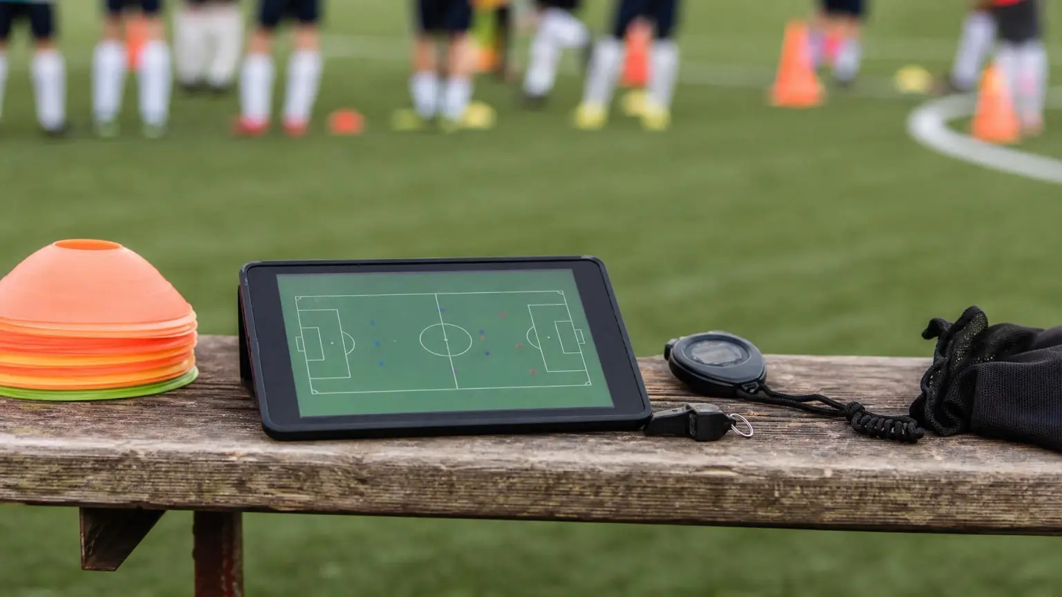 A coach's tablet with a training plan beside a whistle, stopwatch, and cones at the edge of a soccer pitch, representing the best soccer coaching apps