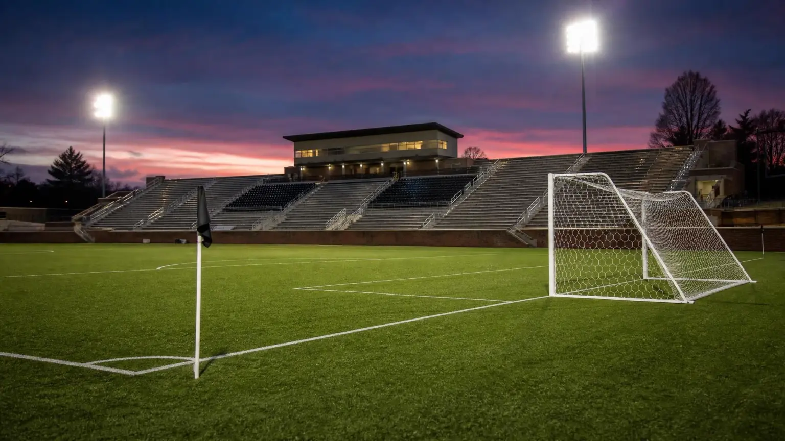An empty collegiate soccer stadium at dusk under floodlights with the goal net and corner flag in the foreground, illustrating how long a college soccer game lasts