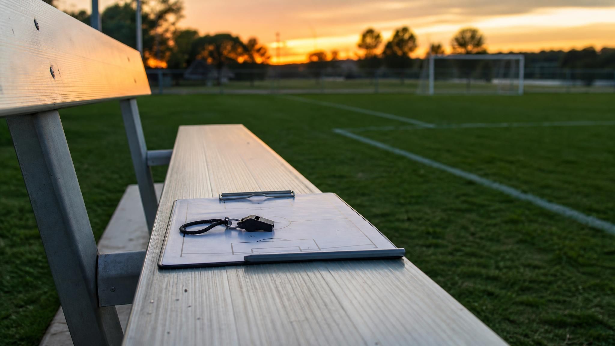 A coach's clipboard and whistle left on an empty bleacher bench beside a youth soccer field at dusk, the quiet moment before a parent talks to their child's soccer coach