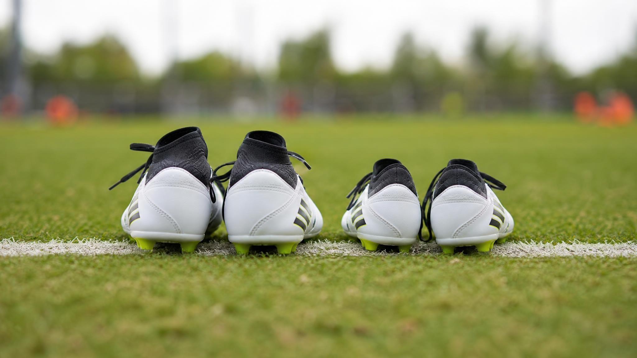 Two pairs of youth soccer cleats of noticeably different sizes side by side on a pitch touchline, illustrating the relative age effect in youth soccer