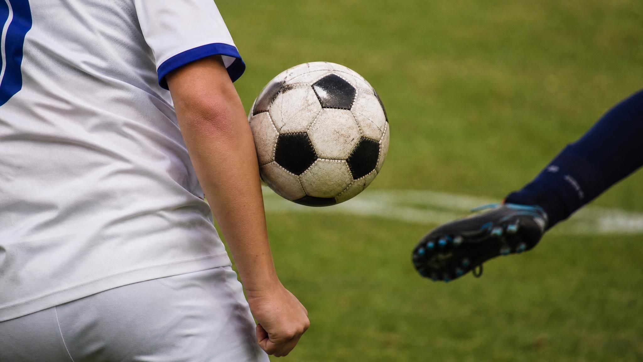 A soccer ball in mid-air inches from a defender's forearm tucked against their side, illustrating the handball rule in soccer