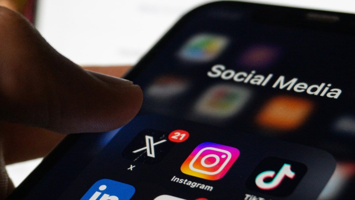 Close-up of a person using a smartphone displaying a folder of social media apps including X, Instagram, and TikTok, under th