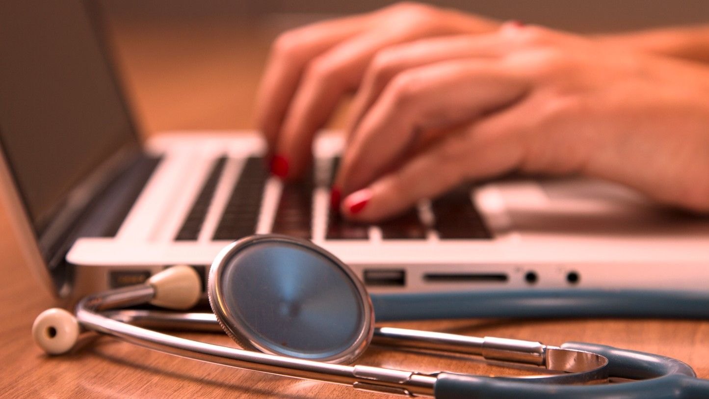 Hands typing on a laptop with a stethoscope on the desk in the foreground