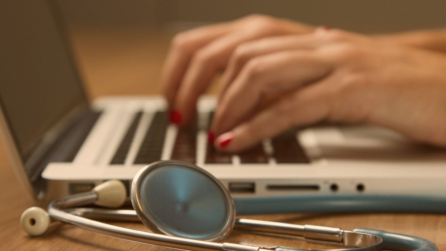 Hands typing on a laptop with a stethoscope on the desk