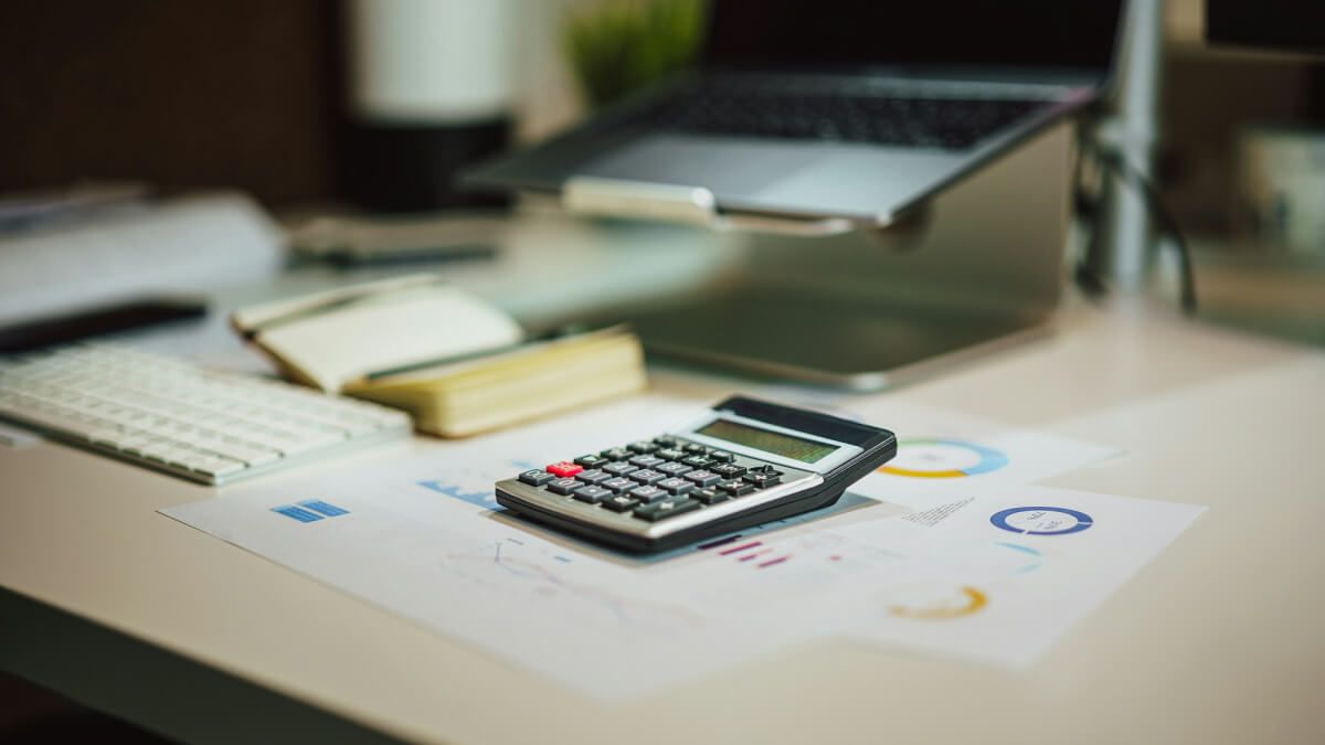 Calculator resting on printed financial charts beside a laptop, representing analysis of inflation and cost-of-living data.