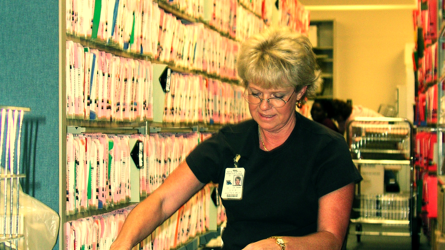 A medical records technician files patient records in front of a wall of health record folders