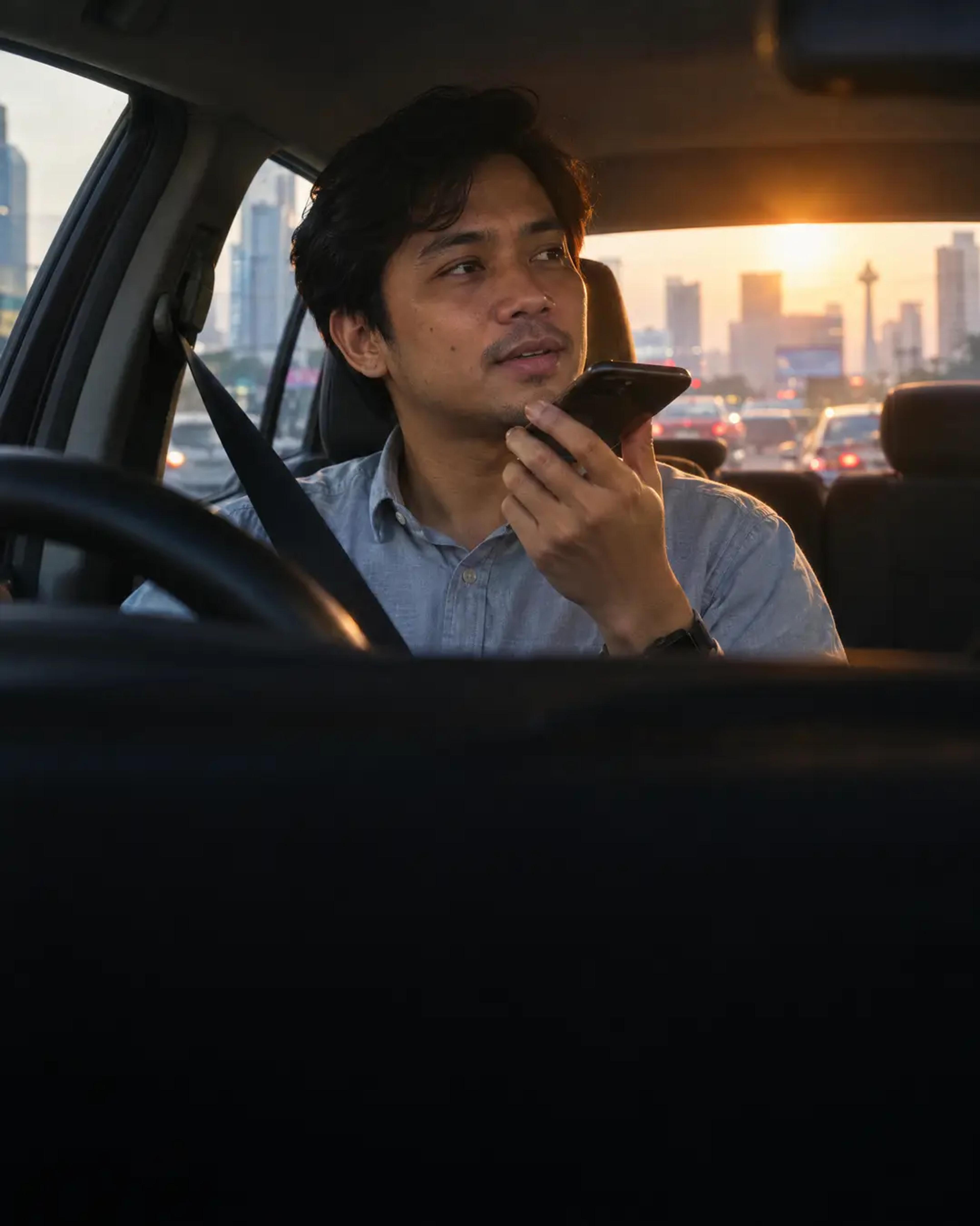 A man in a car seat records a voice note on his phone, city towers at sunrise behind him.