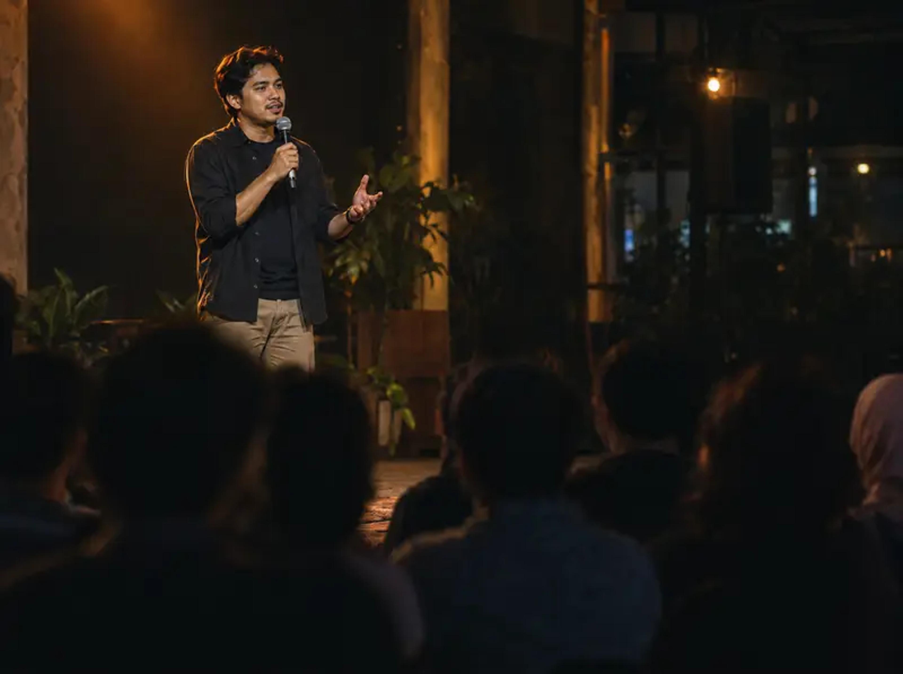A speaker with a microphone tells a story on a small stage to an audience at an evening community event.