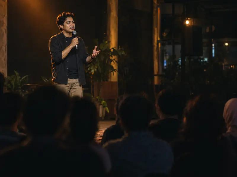 A speaker with a microphone tells a story on a small stage to an audience at an evening community event.