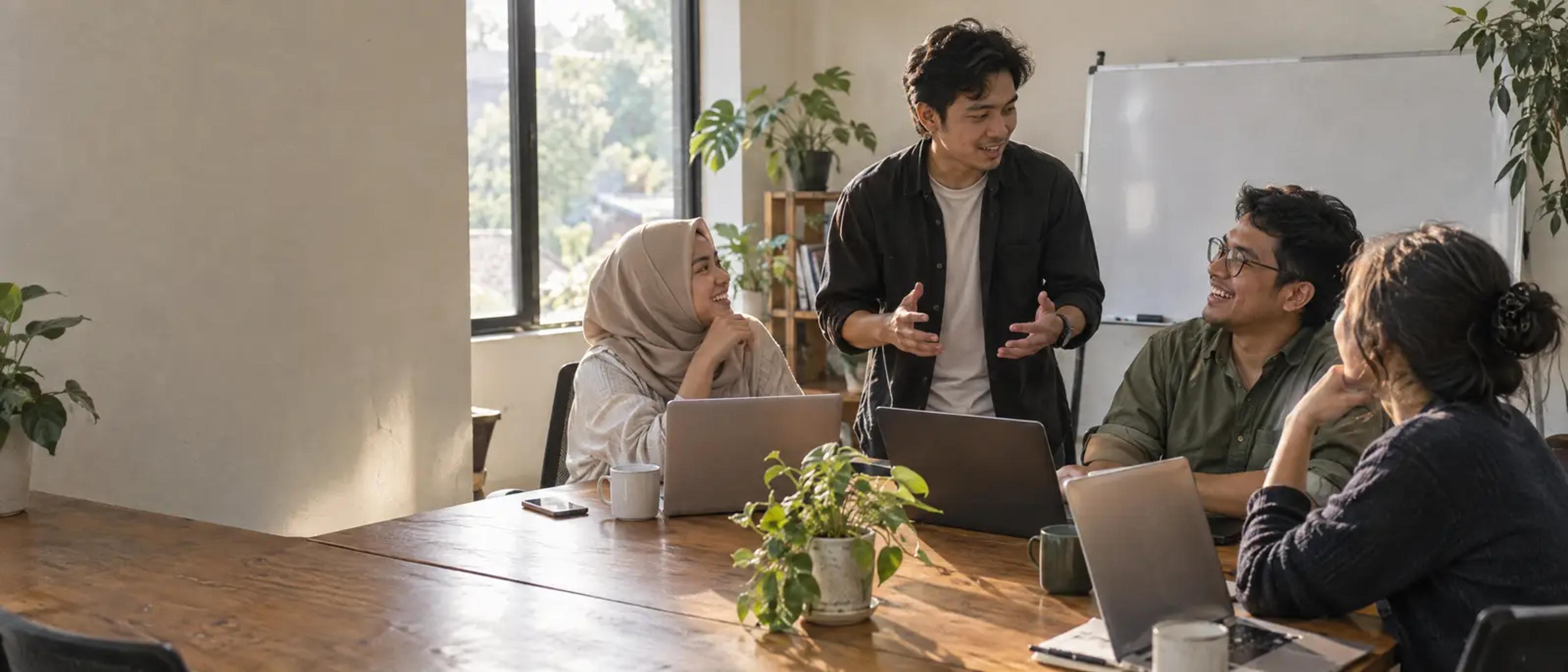 A small team talks over laptops at a long wooden table in a bright office in the afternoon.