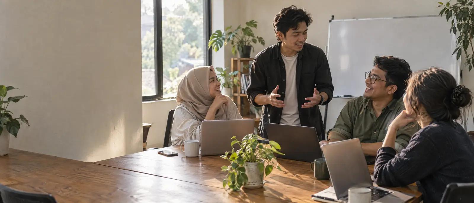 A small team talks over laptops at a long wooden table in a bright office in the afternoon.