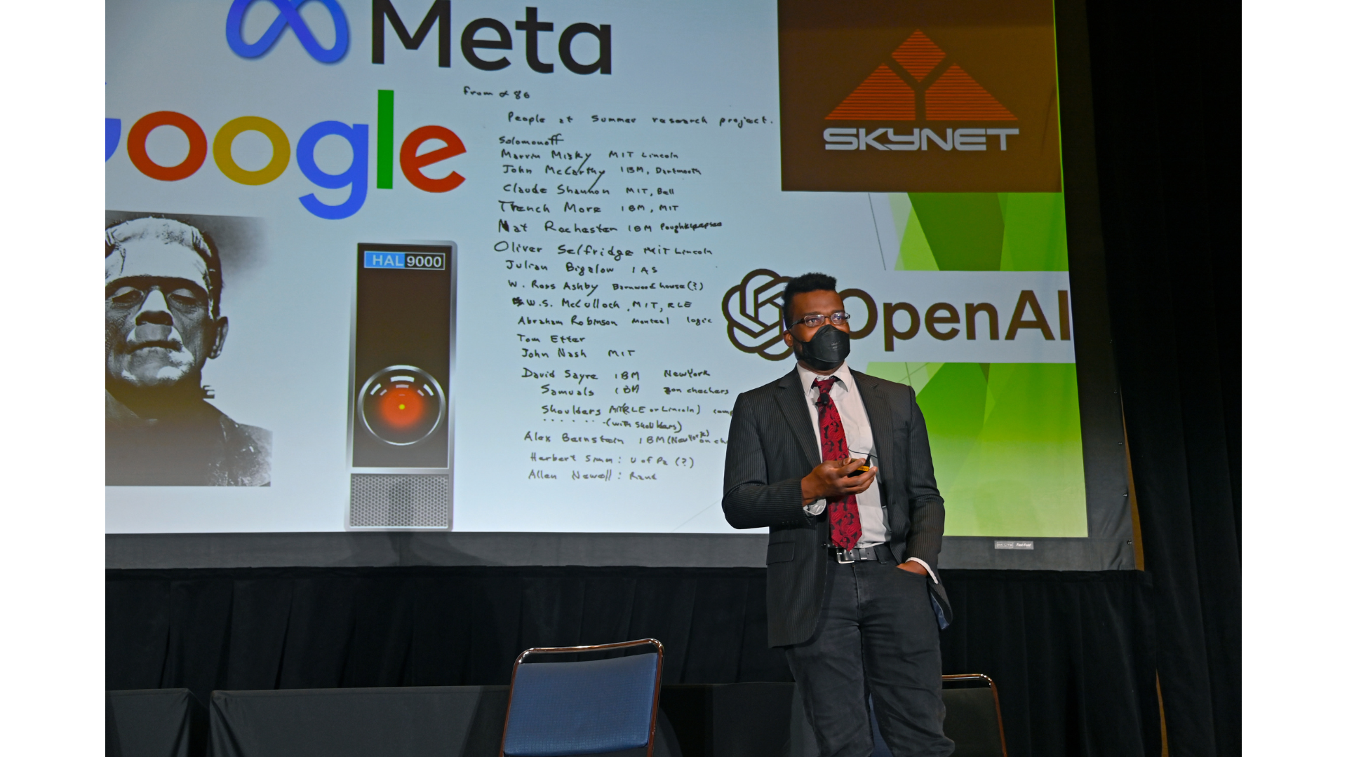 A Black man in a charcoal pinstipe suit jacket, a light grey dress shirt with a red and black Paisley tie, black jeans, black boots, and a black N95 medical mask stands on a stage in front of tables, chairs, and a large screen showing a slide containing images of the meta logo, the skynet logo, the google logo, a headshot of boris karloff as frankenstein's creature, the rectangular black interface with glowing red circle of HAL-9000, the OpenAI logo, and an image of the handwritten list of the attendees of the original 1956 Dartmouth Summer Research Project on Artificial Intelligence (NB: all named attendees are men)