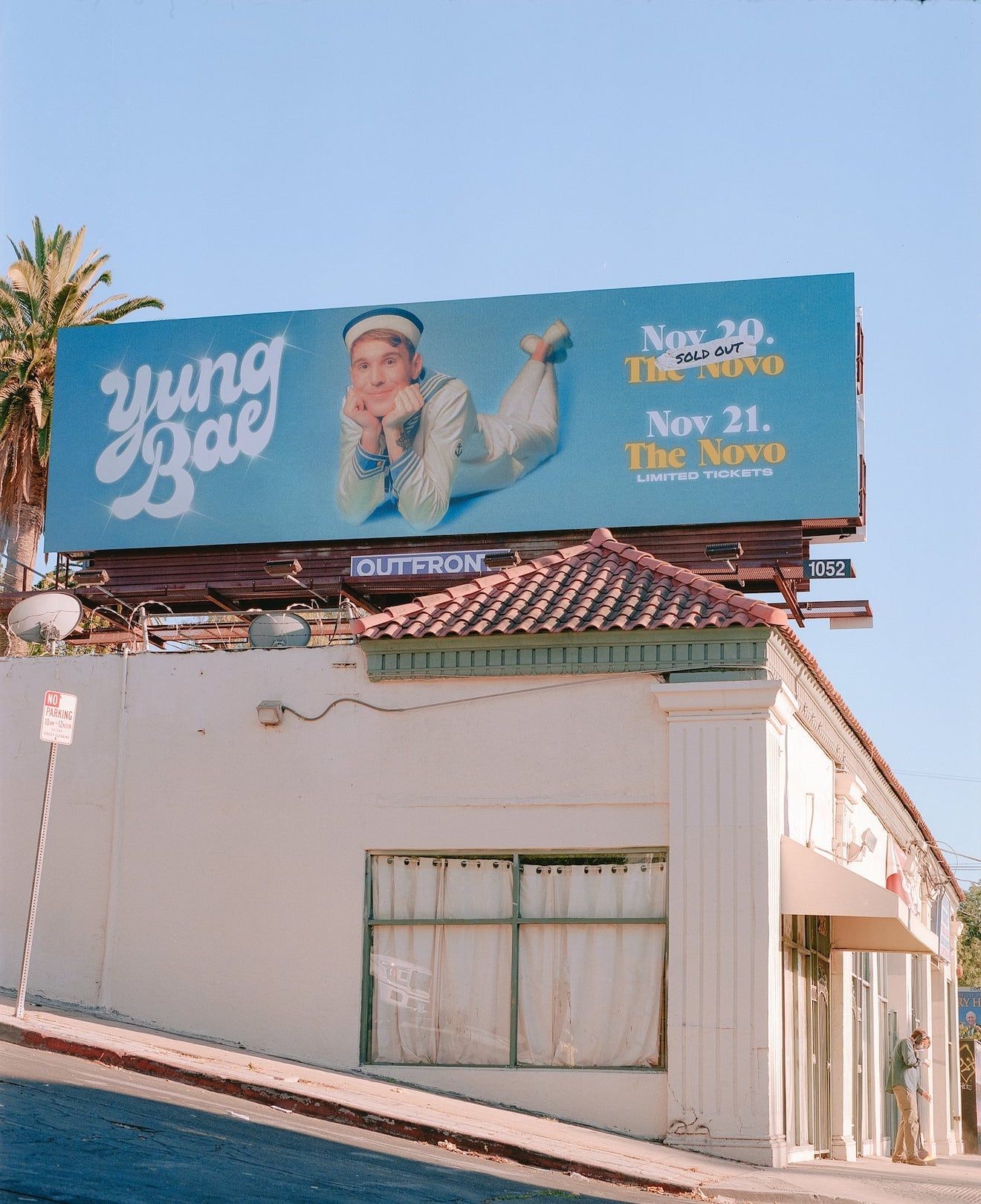 A billboard in LA featuring a smiling Yung Bae in a sailor outfit laying on the ground facing the camera with his head propped up on his hands. To his left is his name drawn in a groovy script and to the right are two dates for upcoming shows.