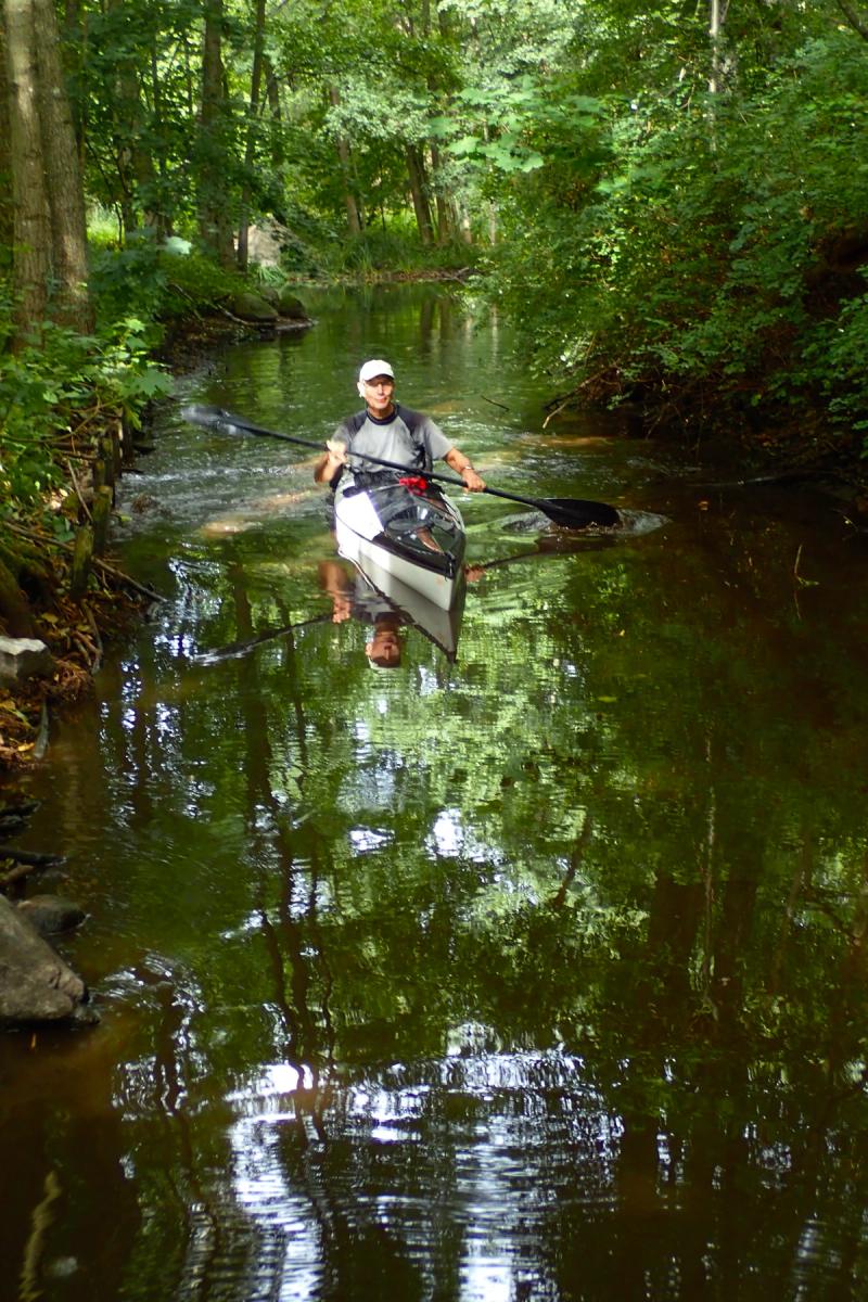 Paddler im Kajak in einem schmalen Bach, in dem sich die Bäume spiegeln.