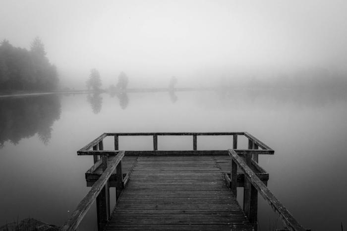 Ein leerer Holzsteg führt hinaus auf stilles, nebelverhangenes Wasser – Schwarzweiß-Aufnahme.