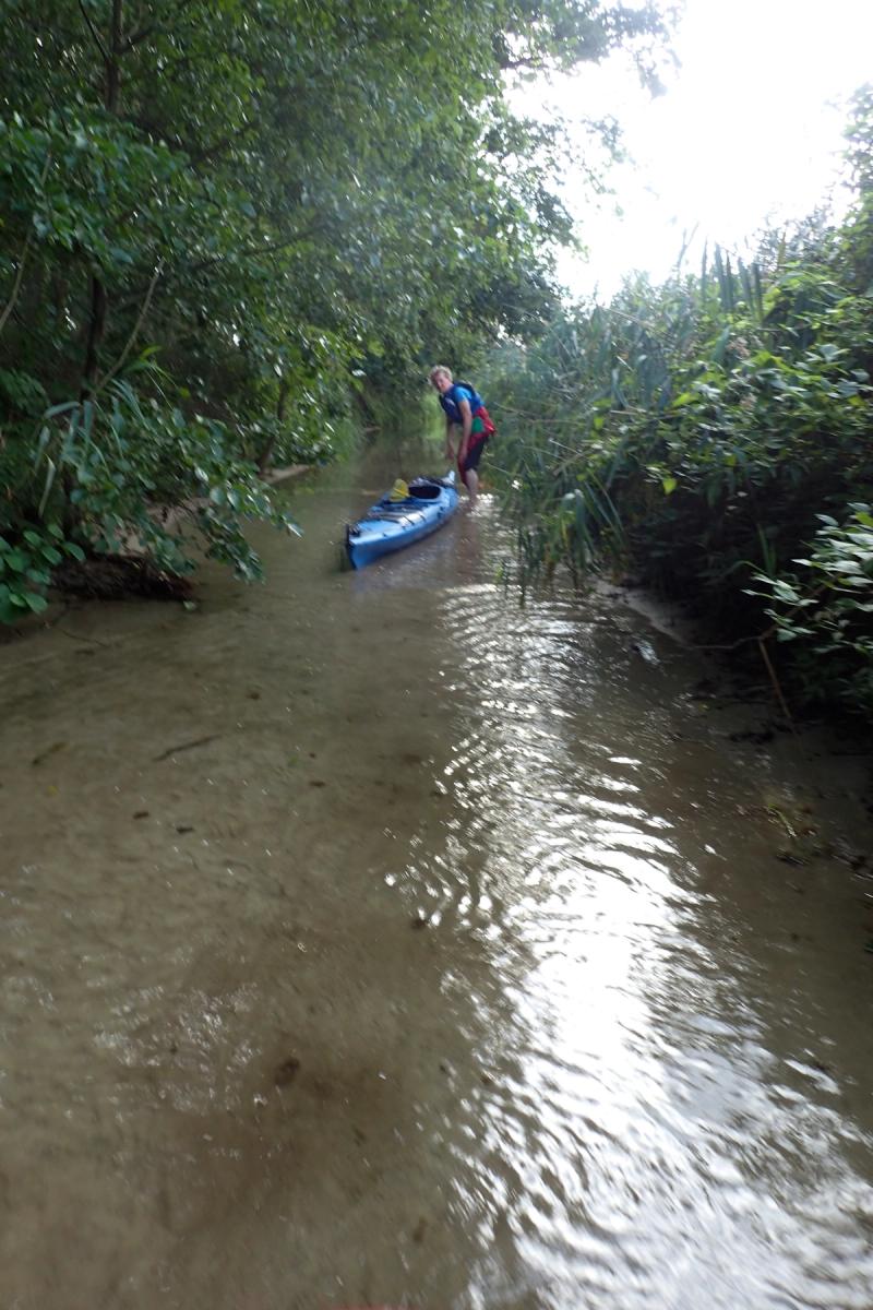 Flacher, schlammiger Kanal: Ein Paddler steht neben seinem Kajak im Wasser.