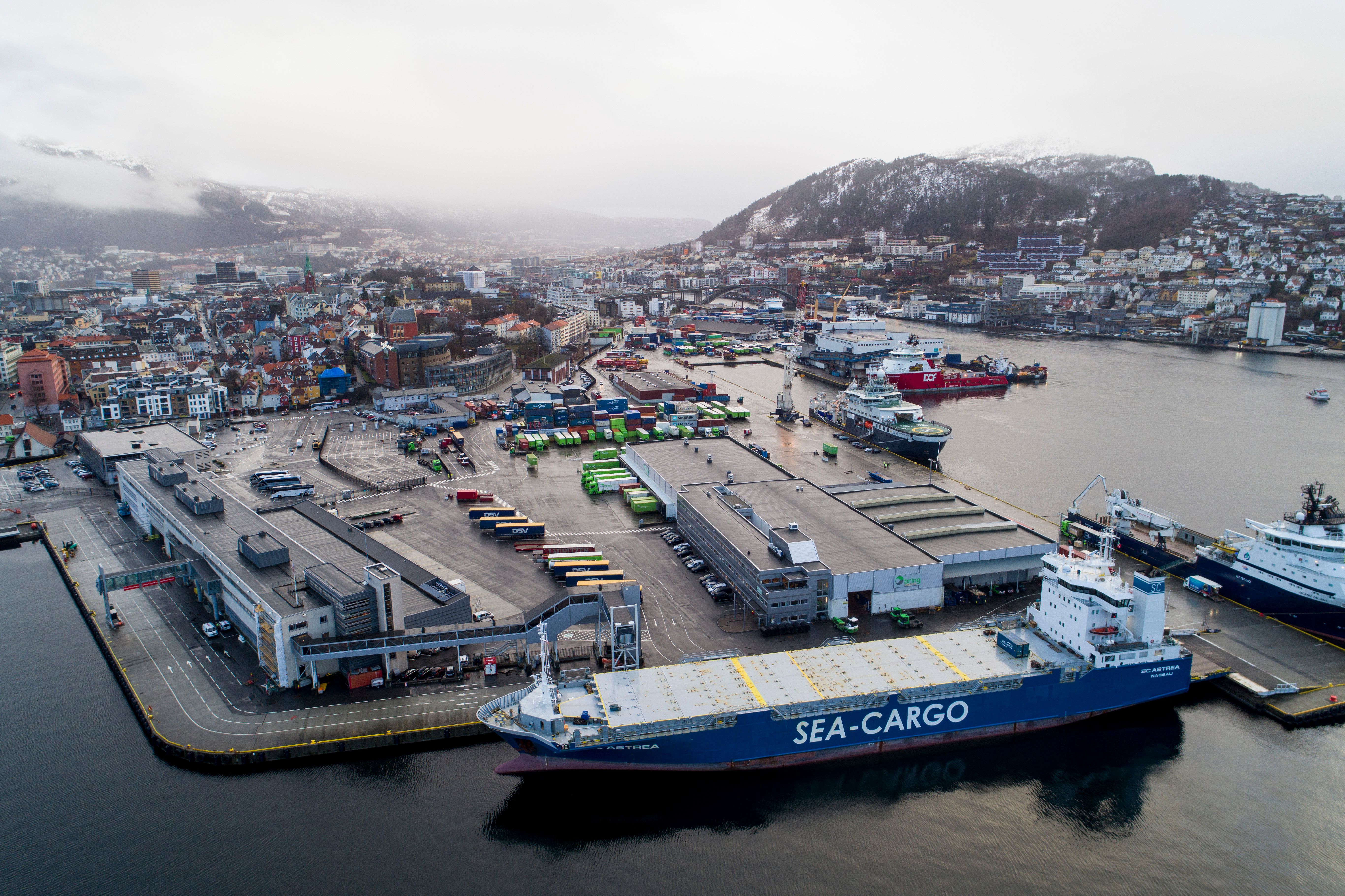 Flyfoto av en travel havn med et SEA-CARGO-skip ved kai, containere, industribygg og en by med snødekte fjell i bakgrunnen.