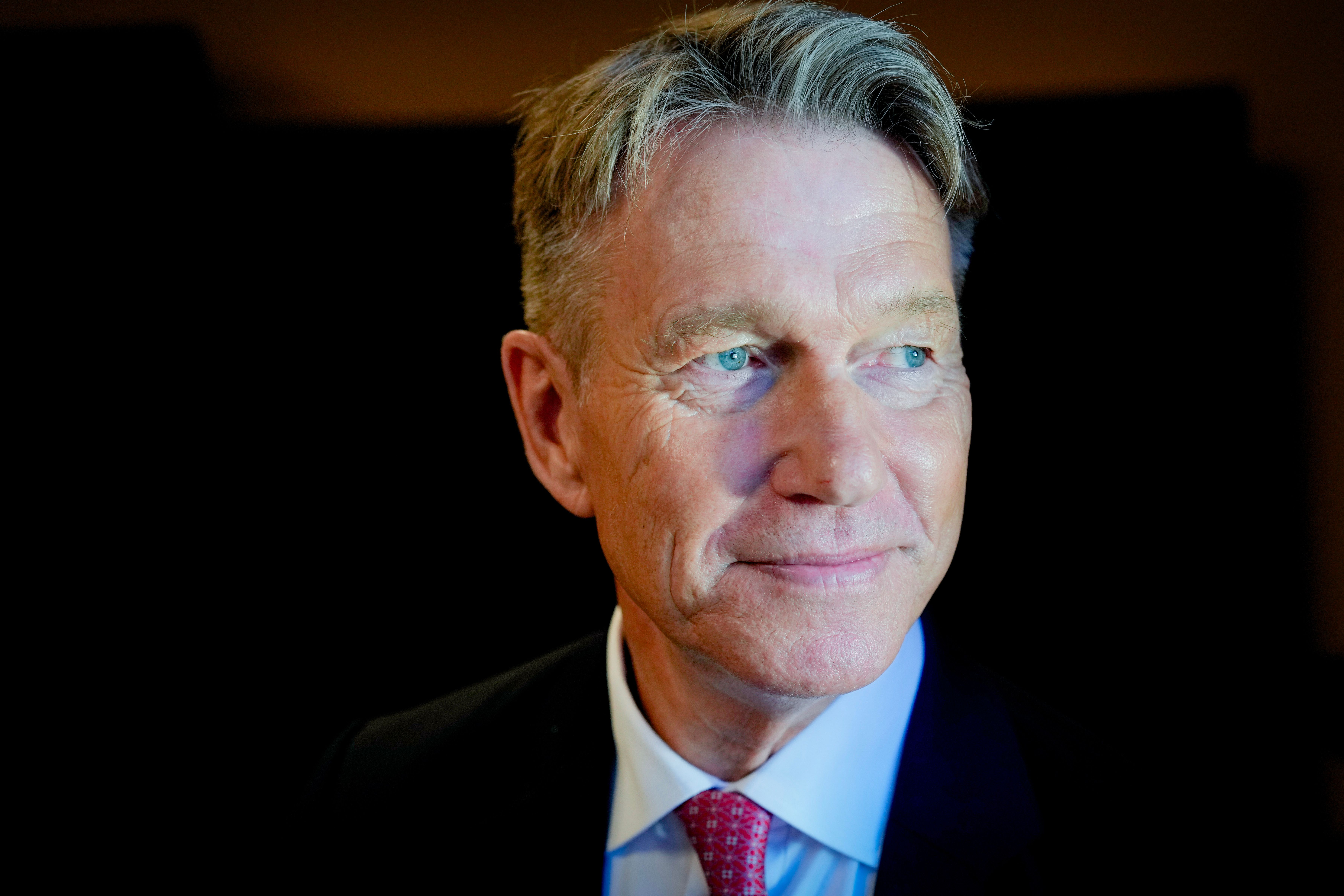 En middelaldrende mann med grått hår og blå øyne, iført dress og rød slips, som smiler og ser mot høyre.