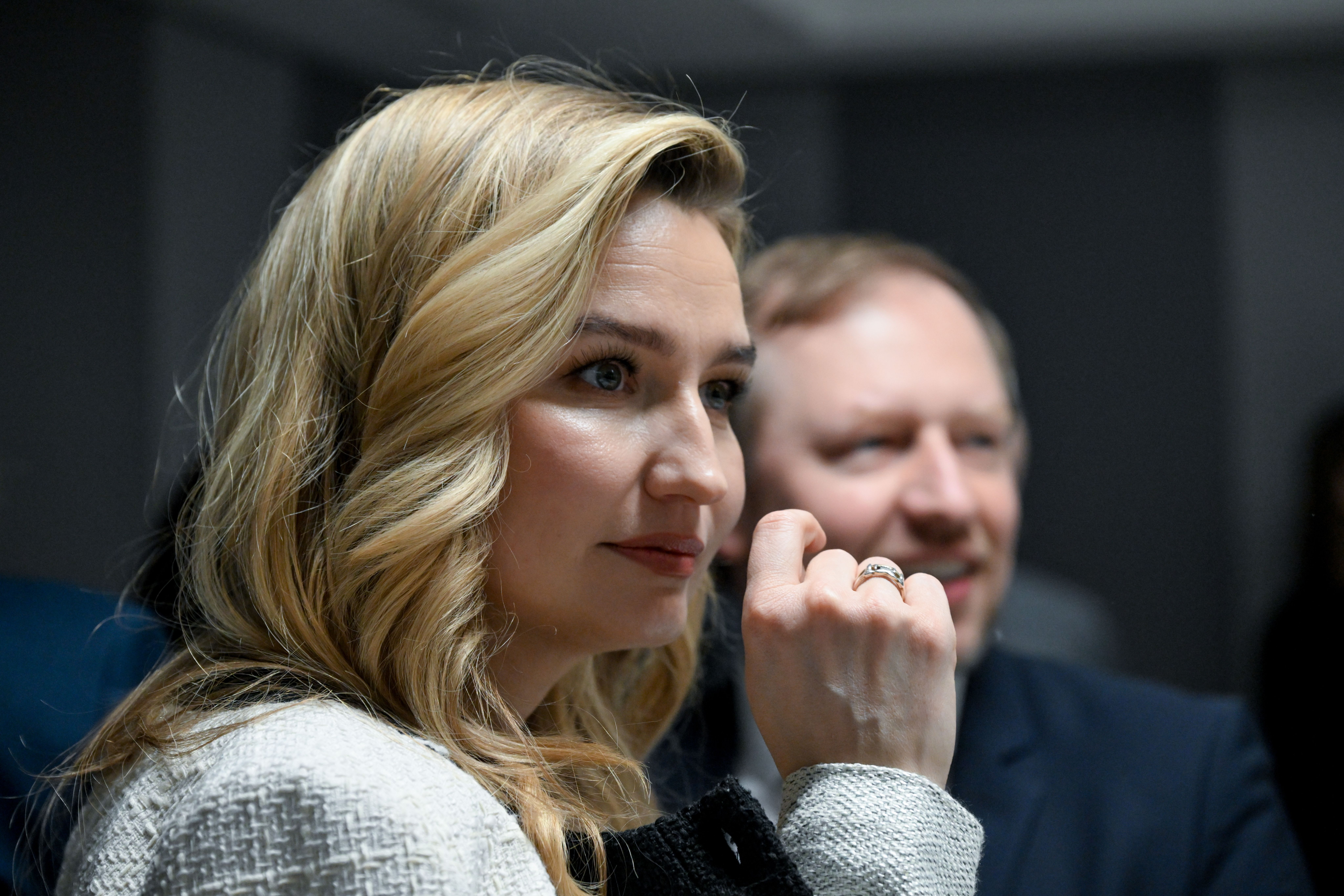 En blond kvinne med hånden under haken ser mot høyre, mens en uklar mann smiler i bakgrunnen.