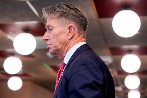 Sideprofil av en mann med grått hår i mørk dress og rød slips, med et alvorlig uttrykk.